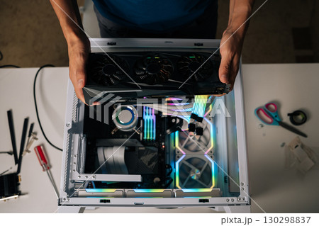 Top view of computer technician installing high-end graphic card into custom gaming pc, showcasing vibrant rgb lighting and advanced water cooling for peak performance enhancement. 130298837
