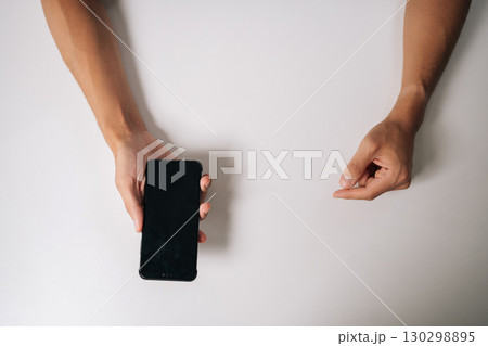 Top view of male hands holding smartphone with blank screen and pointing finger on white background, perfect for showcasing mobile apps or advertisements. Mockup for advertisements or app promotions. Top view of male hands holding smartphone with blank screen and pointing finger on white background, perfect for showcasing mobile apps or advertisements. Mockup for advertisements or app promotions. 130298895