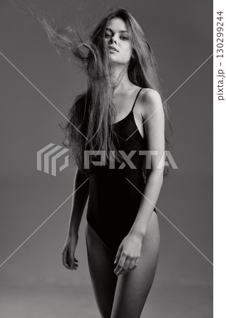 Black and white portrait of young woman in black bodysuit with flowing hair and serious expression on gray background 130299244