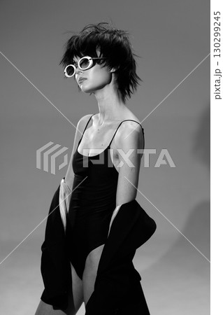 Black and white portrait of a stylish woman wearing sunglasses and a black dress with jacket, posing confidently indoors. 130299245