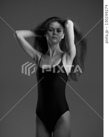 Black and white portrait of a slender woman in a black swimsuit posing confidently against a gray background. Black and white portrait of a slender woman in a black swimsuit posing confidently against a gray background. 130299274