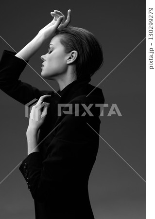Black and white profile of a thoughtful woman in dark clothing with hand on forehead against a plain dark background. 130299279