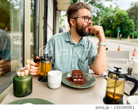 Bearded man sitting at outdoor cafe table looking to side. Modern lifestyle, urban leisure and relaxed atmosphere of everyday social environment. 130300061
