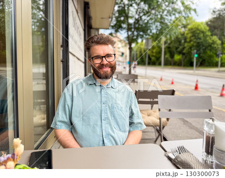 Man with beard smiling at outdoor cafe table on city street. Lifestyle moment of leisure, authenticity and social connection in modern urban environment. 130300073