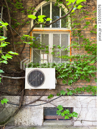 Arched window with ivy and air conditioning unit on brick wall. Cultural architecture combined with modern technology and natural greenery in urban environment. Arched window with ivy and air conditioning unit on brick wall. Cultural architecture combined with modern technology and natural greenery in urban environment. 130300076