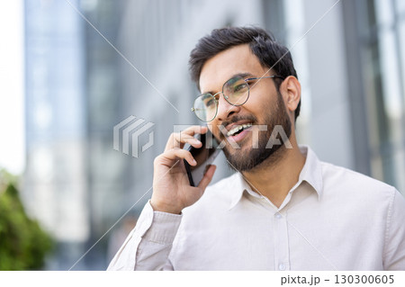 Portrait of a smiling young businessman wearing glasses, speaking on his mobile phone in front of an office building, enjoying a pleasant conversation Portrait of a smiling young businessman wearing glasses, speaking on his mobile phone in front of an office building, enjoying a pleasant conversation 130300605