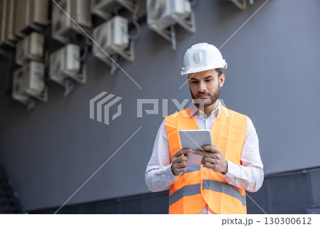 Building maintenance technician wearing safety helmet and vest, using digital tablet and inspecting air conditioning units on building exterior 130300612