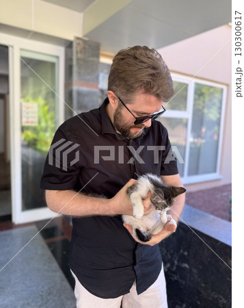 Man in sunglasses smiling and holding small kitten outdoors. Companionship affection, and emotional bond between human and animal. 130300617