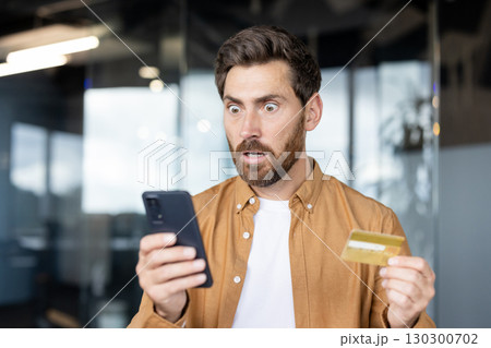 A shocked man looks at his phone screen while holding a credit card, possibly reacting to an unexpected charge or financial issue. 130300702