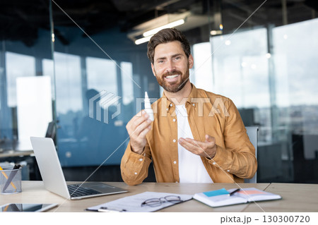 Man sitting at a modern office desk, presenting a small bottle of nose drops with a smile. Man sitting at a modern office desk, presenting a small bottle of nose drops with a smile. 130300720
