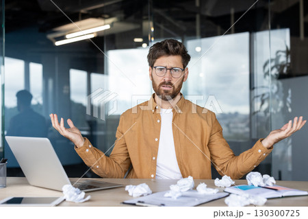 Office worker shrugging in confusion, surrounded by crumpled papers on the desk, grappling with writer's block and struggling to complete the project at hand 130300725