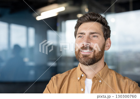 Smiling man wearing a casual shirt standing in a bright contemporary office space with large windows. The professional setting and his positive expression reflect confidence, success 130300760
