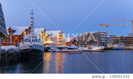 Evening scene at Tromso harbor featuring docked boats under a snowy landscape. Warm lights from red and yellow buildings reflect on the water, creating a serene winter atmosphere 130301242