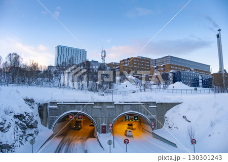 Vehicles navigate a snowy tunnel in Tromso, Norway, surrounded by a winter landscape and urban architecture. Snow-covered trees and buildings frame the scene in serene twilight Vehicles navigate a snowy tunnel in Tromso, Norway, surrounded by a winter landscape and urban architecture. Snow-covered trees and buildings frame the scene in serene twilight 130301243
