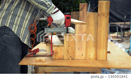 Worker in gloves using nail gun to fasten wooden planks into step-shaped structure during carpentry 130301841
