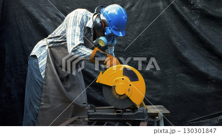 Worker using yellow metal cutting saw while wearing blue hard hat gloves apron ear protection and respirator mask Worker using yellow metal cutting saw while wearing blue hard hat gloves apron ear protection and respirator mask 130301847