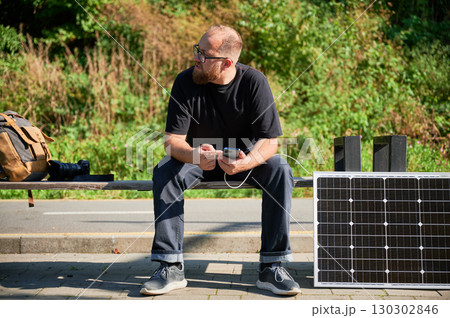 Bearded man using smartphone connected to photovoltaic solar panel. Integration of sustainable renewable energy into everyday life, demonstrating practical use of solar power for charging devices. 130302846