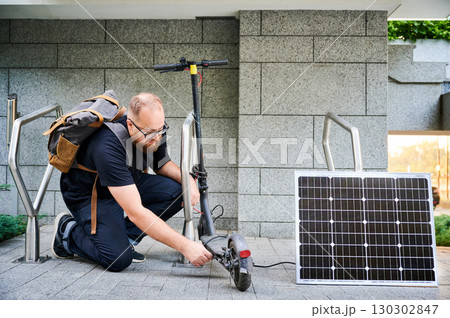 Man connects solar panel to electric scooter for charge, highlighting practical application of sustainable renewable energy. Concept of integration of eco-friendly technology in urban transportation. Man connects solar panel to electric scooter for charge, highlighting practical application of sustainable renewable energy. Concept of integration of eco-friendly technology in urban transportation. 130302847