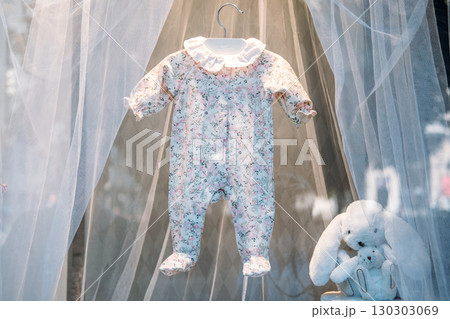 Floral baby romper hanging on a white hanger inside a softly lit display with sheer curtains and plush toys. Vintage childhood, boutique nursery style, delicate textures, early parenthood Floral baby romper hanging on a white hanger inside a softly lit display with sheer curtains and plush toys. Vintage childhood, boutique nursery style, delicate textures, early parenthood 130303069