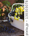 Heap of ripe grape in white basket on wooden bench. Autumn harvest. Autumn fruits. Bunch of juicy black and white grape in sunlight. Autumn agriculture. September nature. Healthy organic grapes.  130303078