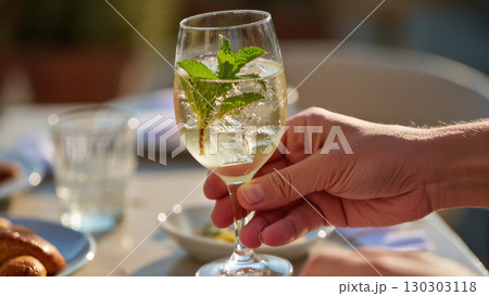 Hand holding glass of Hugo cocktail with mint and ice cubes on summer table outdoors, refreshing elderflower drink 130303118