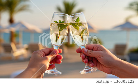 Two people clinking glasses of Hugo cocktail with mint and ice at seaside resort, refreshing summer toast moment 130303122