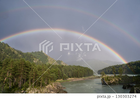 Double rainbow over serene river valley with lush green trees and overcast sky 130303274