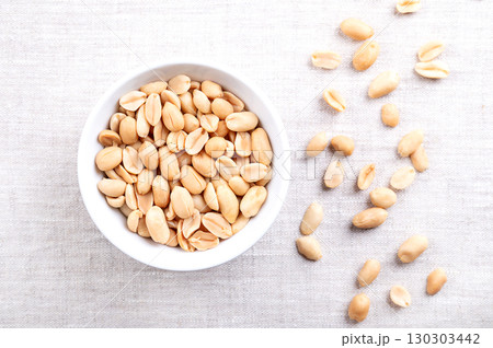 Roasted peanuts in a white bowl on linen fabric. Ready-to-eat crunchy snack food made from fruits of Arachis hypogaea. Light golden snack nuts with natural, nutty aroma, and firm, crunchy texture. 130303442