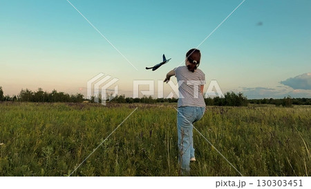 Girl throwing toy plane in the field, Child flying a model airplane outdoors, Summer evening with a toy glider, Nature and childhood fun, Playing in the countryside, Kid launches foam airplane 130303451