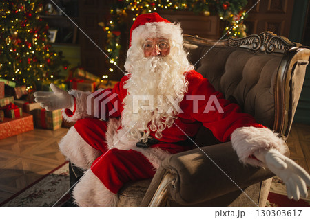 Merry Christmas. Happy Santa Claus sitting in chair in festive living room with fireplace decorations Christmas tree. Santa in cozy home on Christmas eve. Winter New Year Holiday Celebrations Merry Christmas. Happy Santa Claus sitting in chair in festive living room with fireplace decorations Christmas tree. Santa in cozy home on Christmas eve. Winter New Year Holiday Celebrations 130303617