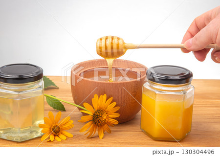 Honey dripping from a wooden dipper into a bowl with jars of honey and flowers nearby 130304496