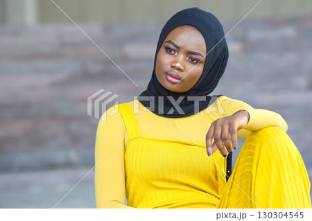 Elegant African woman in yellow attire with a black hijab poses thoughtfully outdoors 130304545