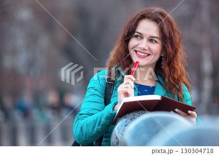 Young woman enjoys writing in her notebook while standing outdoors in a park on a sunny day Young woman enjoys writing in her notebook while standing outdoors in a park on a sunny day 130304718