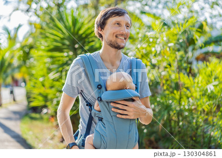 Young father walks in nature with his baby son nestled close in a parent-facing ergonomic baby carrier. Bonding, warmth, and modern parenting concept in a peaceful outdoor setting 130304861