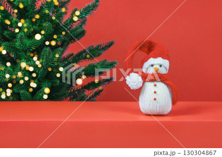 Christmas red background with knitted snowman in red hat and Christmas tree with golden lights and space for text, greetings, promotions, and discounts. Christmas and New Year card. Christmas red background with knitted snowman in red hat and Christmas tree with golden lights and space for text, greetings, promotions, and discounts. Christmas and New Year card. 130304867