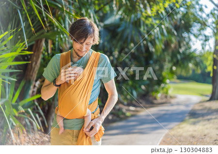 Father holding his little baby in a yellow sling in the park. Warm and loving family moment. Babywearing, fatherhood, and parent-child bonding concept 130304883