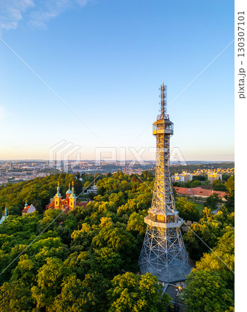 Petrin Lookout Tower rises majestically, surrounded by lush greenery and historic buildings. The sunrise bathes Prague in warm hues, enhancing the scenic beauty of the city. 130307101