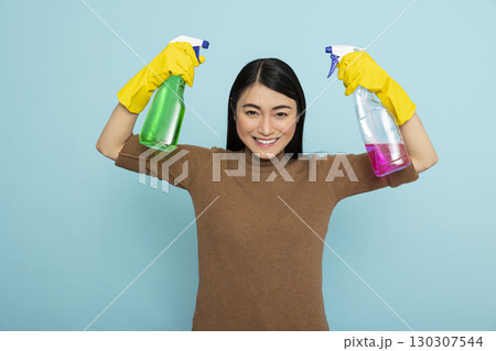 Cheerful female janitor flexing biceps with pink and green liquid solutions, excited to provide cleaning services. Happy asian housekeeper holding disinfectant spray bottles and looking at camera. Cheerful female janitor flexing biceps with pink and green liquid solutions, excited to provide cleaning services. Happy asian housekeeper holding disinfectant spray bottles and looking at camera. 130307544