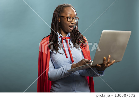 Shocked african american woman in a red cape stares at her laptop with wide eyes. Black female model with expressive reaction conveying excitement and unexpected positive news. Shocked african american woman in a red cape stares at her laptop with wide eyes. Black female model with expressive reaction conveying excitement and unexpected positive news. 130307572