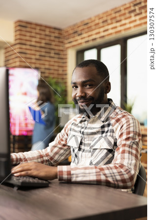 Portrait of confident male employee working on his computer, typing in modern workspace. African American businessman sitting at his desk, using desktop pc and looking at camera in brick wall office. Portrait of confident male employee working on his computer, typing in modern workspace. African American businessman sitting at his desk, using desktop pc and looking at camera in brick wall office. 130307574