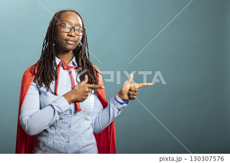 African American woman in red superhero cape gestures to side with index fingers, standing against isolated background with neutral expression. Serious black female individual pointing sideways. 130307576
