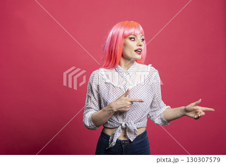 Excited woman with pink wig stands isolated on pink background, smiling and pointing finger towards blank copy space. Energetic and vibrant model presenting area for advertisement or promotional 130307579