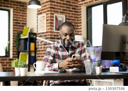 Portrait of happy african american freelancer sitting and using smartphone at modern home office desk. Self employed black man resting from work, networking online using mobile device. 130307586