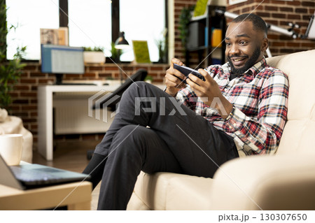 Cheerful man sitting on couch, smiling brightly at cellphone screen while playing mobile game. African american remote worker enjoying fun break from laptop tasks in cozy home setting. Cheerful man sitting on couch, smiling brightly at cellphone screen while playing mobile game. African american remote worker enjoying fun break from laptop tasks in cozy home setting. 130307650