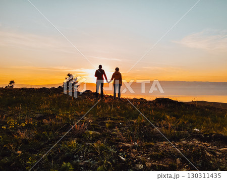 couple holding hands at sunset on hillside overlooking tranquil lake in early evening. 130311435