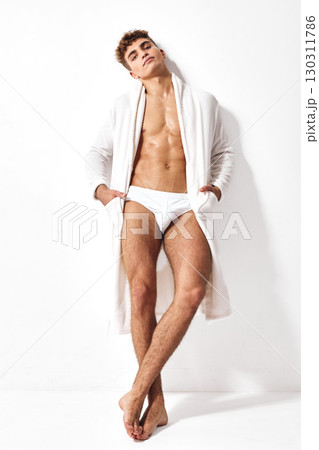 Confident young man posing in a white bathrobe and briefs, relaxing against a clean white background. Fashion and lifestyle concept. 130311786