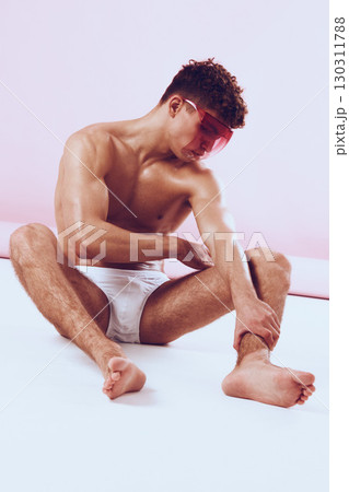 Stylish young man with curly hair sitting on the floor, wearing pink sunglasses and white underwear, posing against a soft pink background. Fashion and self-expression concept. Stylish young man with curly hair sitting on the floor, wearing pink sunglasses and white underwear, posing against a soft pink background. Fashion and self-expression concept. 130311788