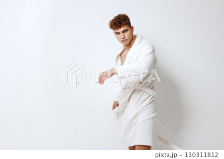 Confident young man in a white bathrobe standing against a plain light background. Fashion and lifestyle concept showcasing relaxation and self-care. Confident young man in a white bathrobe standing against a plain light background. Fashion and lifestyle concept showcasing relaxation and self-care. 130311812