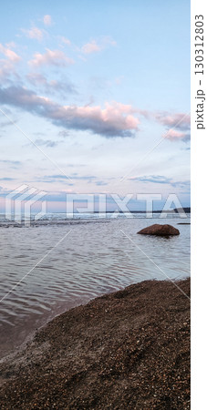 Spring landscape. Panoramic view of beautiful dawn on bay. Cumulus clouds over water in bright light. Ice, snow and rocks on coastline. Rising rays of sun are reflected in sea. Vertical photo. 130312803