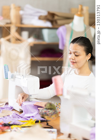 Attentive woman tailor sewing textile using sewing machine sitting at workplace in fashionable sewing atelier Attentive woman tailor sewing textile using sewing machine sitting at workplace in fashionable sewing atelier 130313791